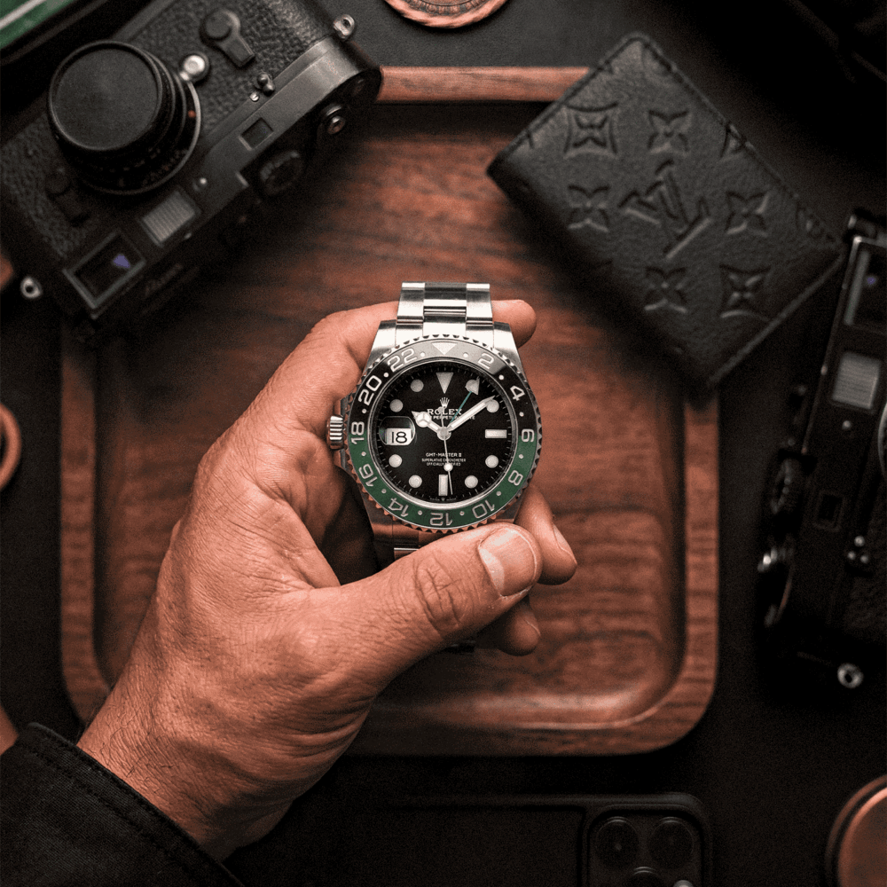 Rolex GMT-Master II Left-Hand "Sprite" held above a table with a valet tray, a film camera, a Louis Vuitton card holder and an iPhone 16 pro max